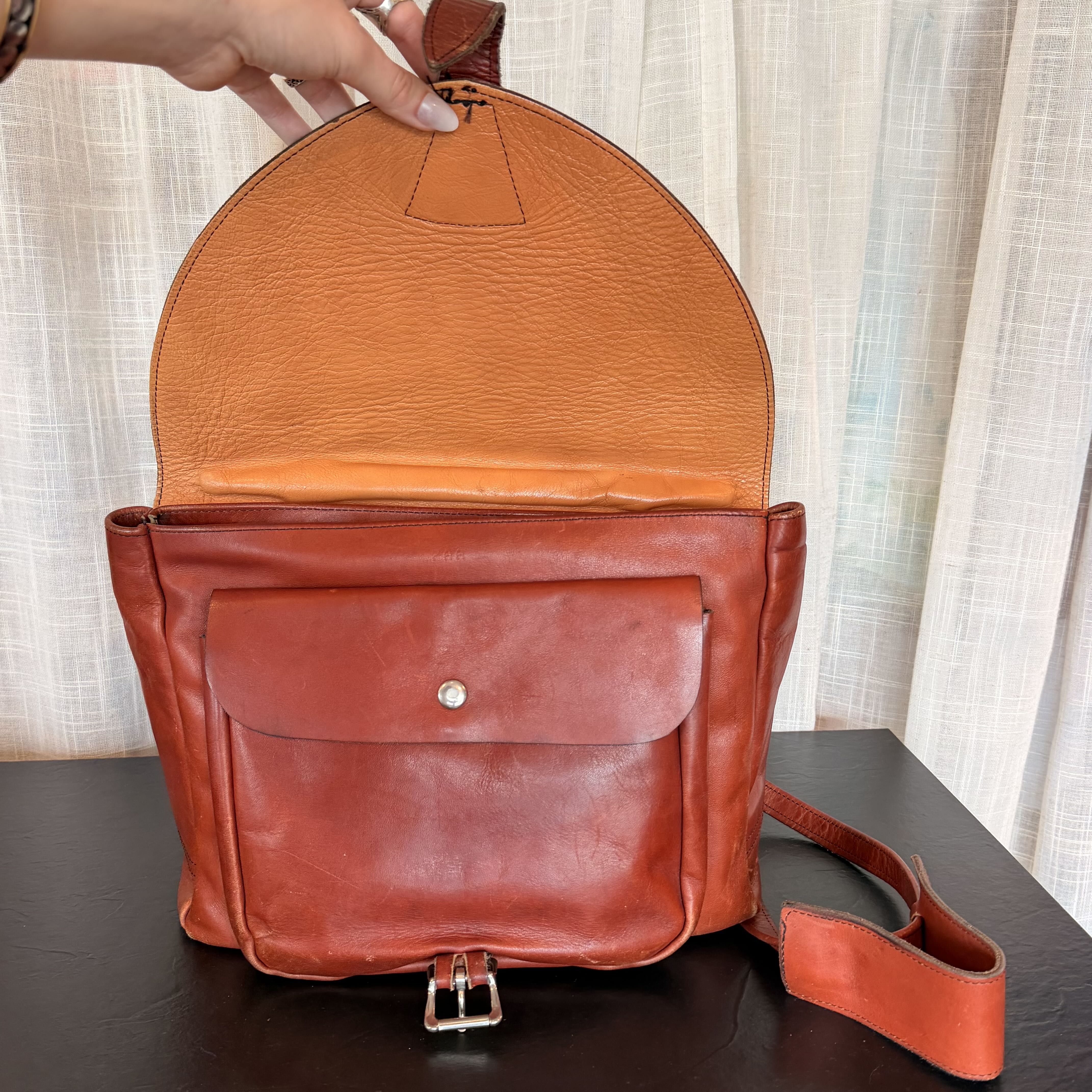 A leather messenger bag in a reddish-brown color, shown open with a hand holding the top handle. The bag features a front pocket with a snap button, a silver buckle, and a matching leather shoulder strap, set against a background of sheer curtains and a dark table.