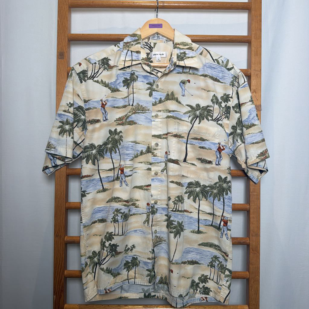 A short-sleeve camp collar button-up shirt with a tropical beach and golf course print, hanging on a wooden hanger on a wooden ladder against a light gray wall.