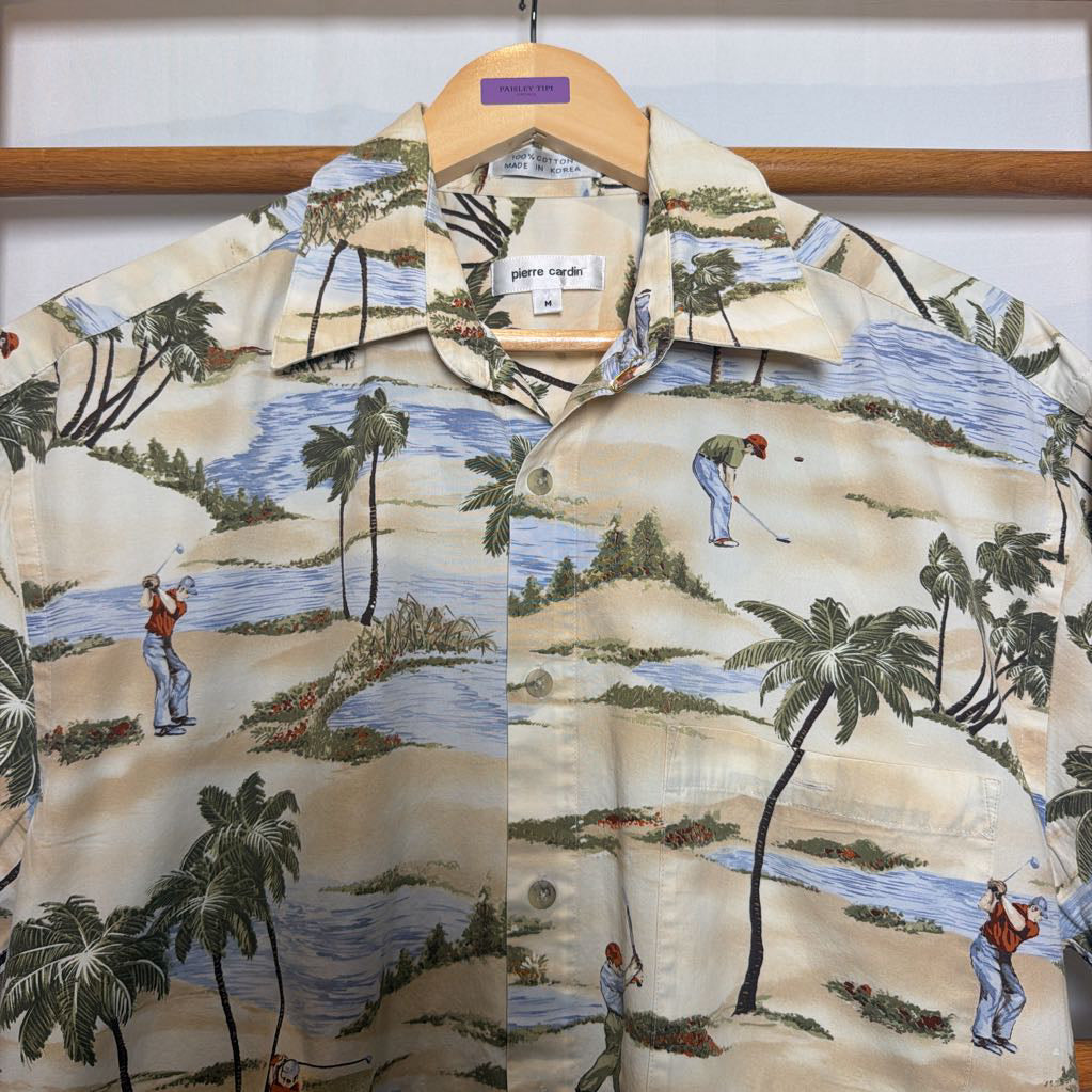 A short-sleeve button-up camp shirt with a golf course landscape print, hanging on a wooden hanger. The shirt features a beige background with illustrations of palm trees, sand dunes, water, and golfers.