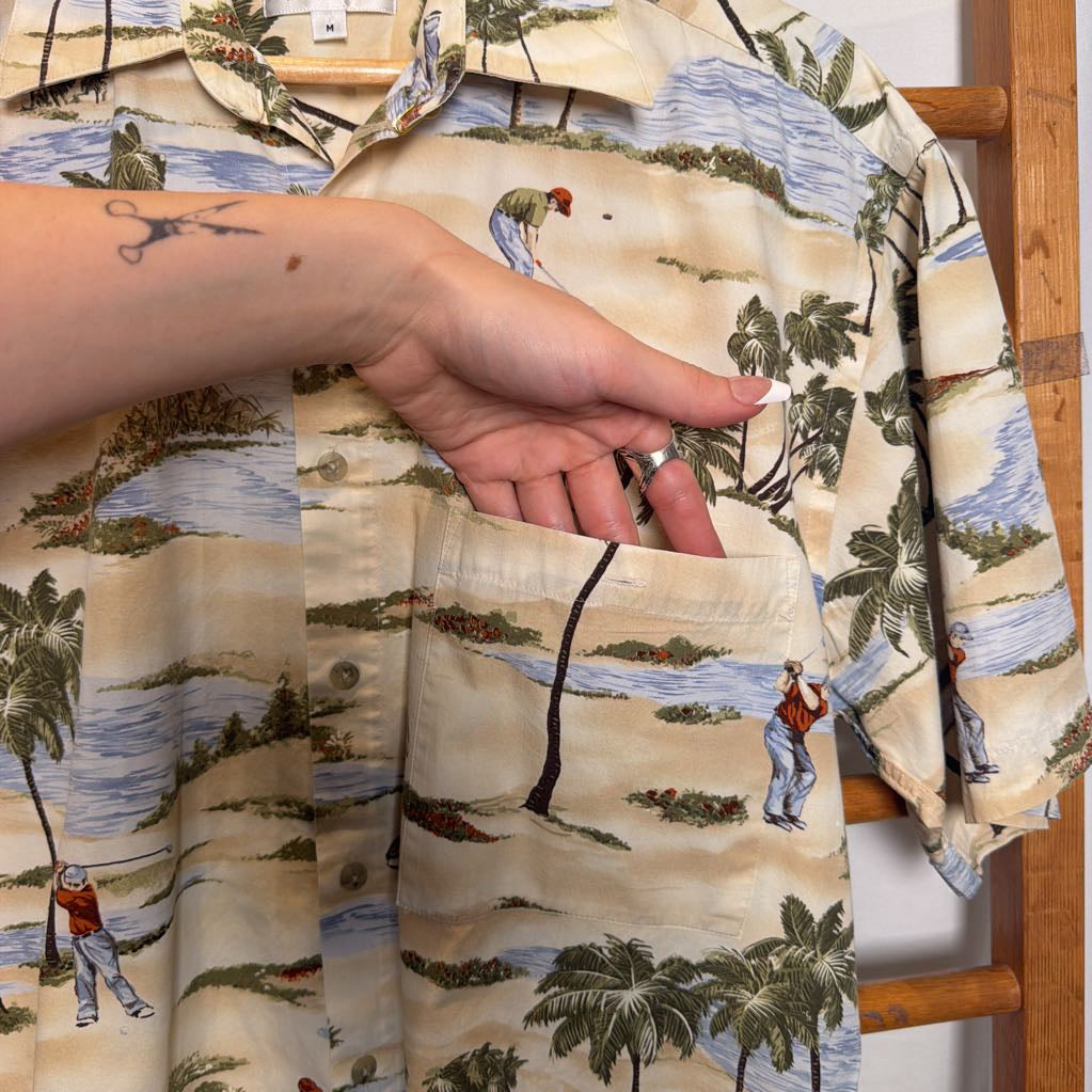 A close-up of a short-sleeve button-up shirt with a tropical beach and golf course print, featuring a hand holding the pocket to show the fabric detail.