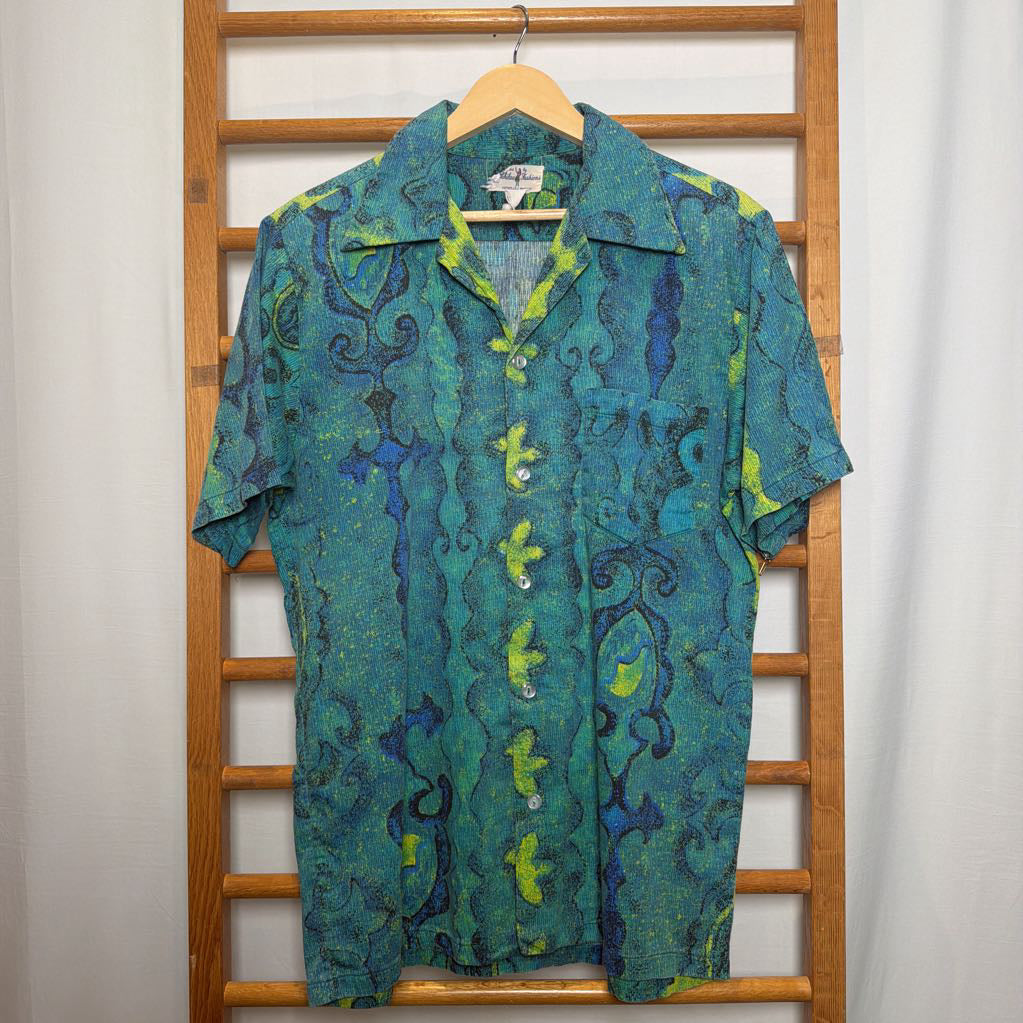 A short-sleeve button-up camp shirt with a green and blue abstract pattern, hanging on a wooden hanger against a wooden slatted backdrop.