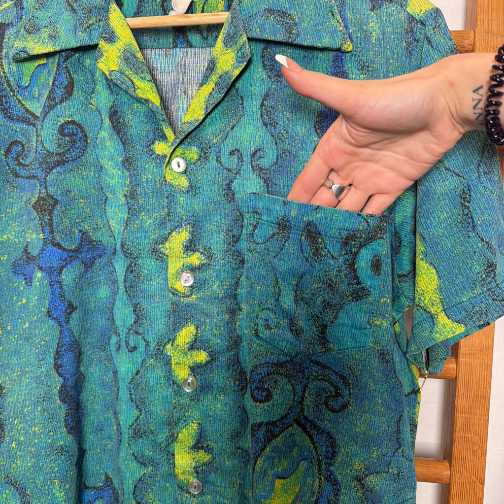 A short-sleeve button-up shirt with a textured, abstract wavy pattern in teal, green, and dark blue, held up on a wooden hanger with a hand pointing to the chest pocket.