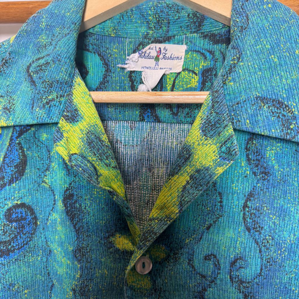 A close-up of a vintage collared shirt jacket by Anita Fashions, featuring a vibrant blue, green, and yellow abstract pattern on a textured fabric, hanging on a wooden hanger.
