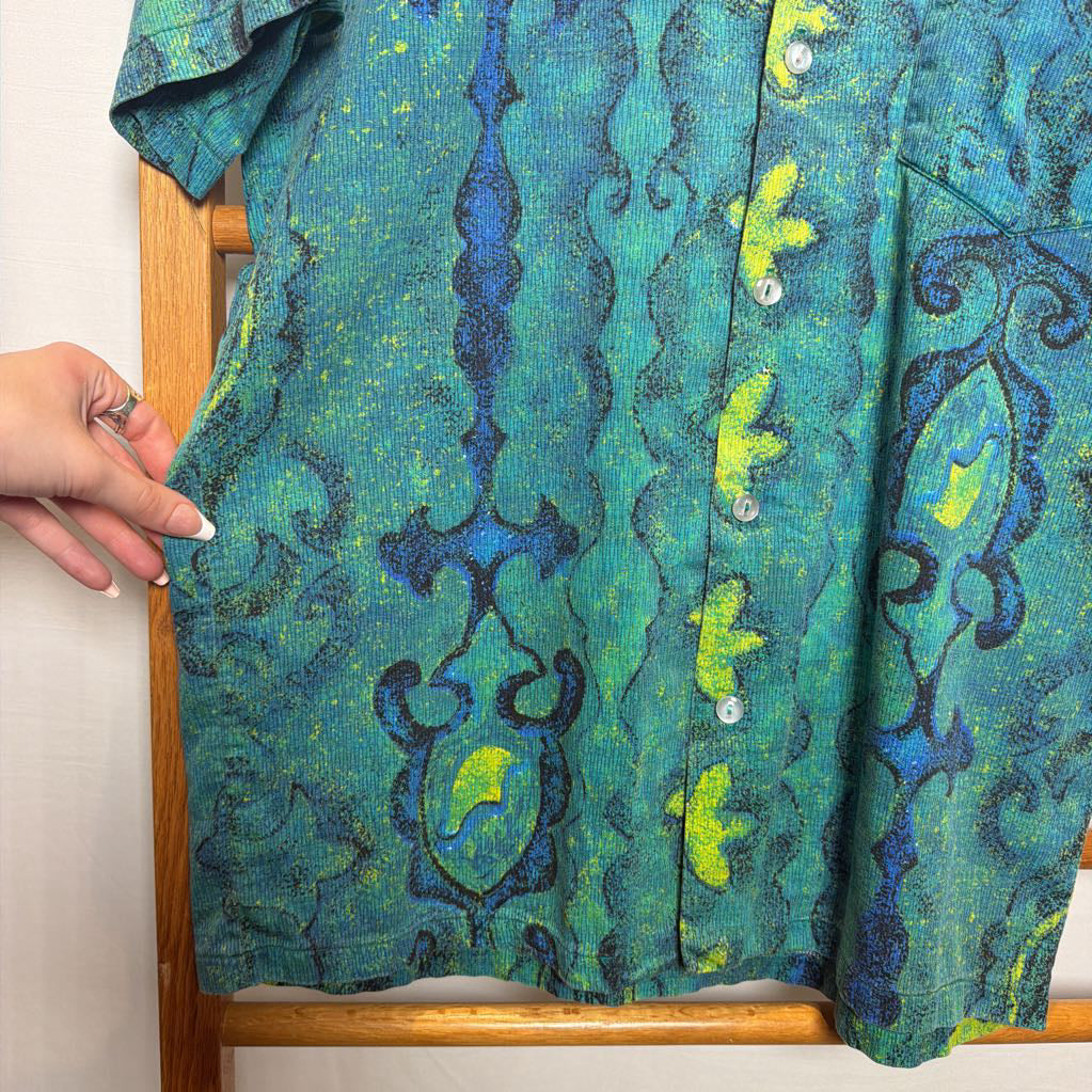 A short-sleeve button-up shirt with a vibrant abstract pattern in teal, green, and blue, held up by a wooden hanger with a hand visible on the left side.