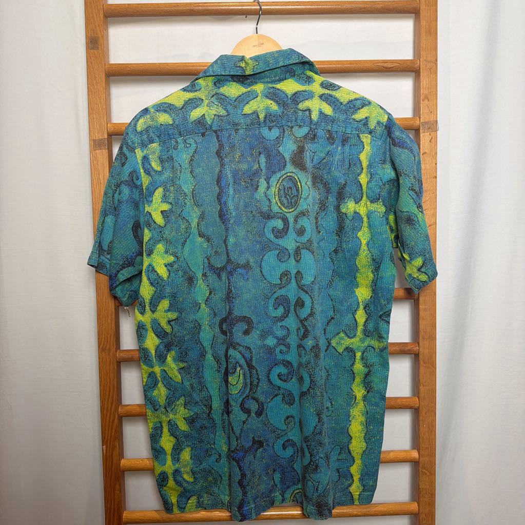 A short-sleeve button-up shirt with a blue and green tribal-style pattern, hanging on a wooden hanger from a wooden clothes rack against a white curtain backdrop.