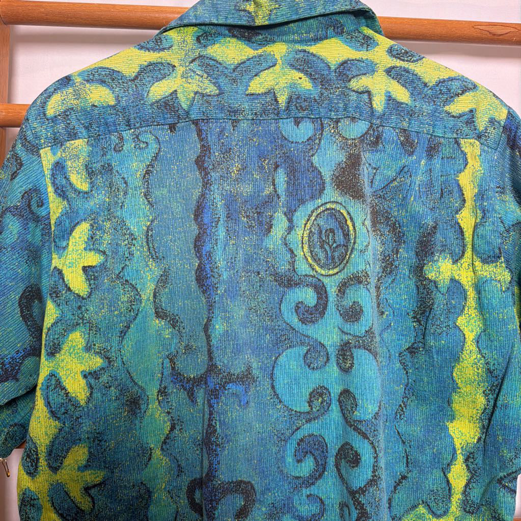 A short-sleeve button-up shirt with a vibrant blue and yellow abstract pattern, hanging on a wooden hanger against a light background.