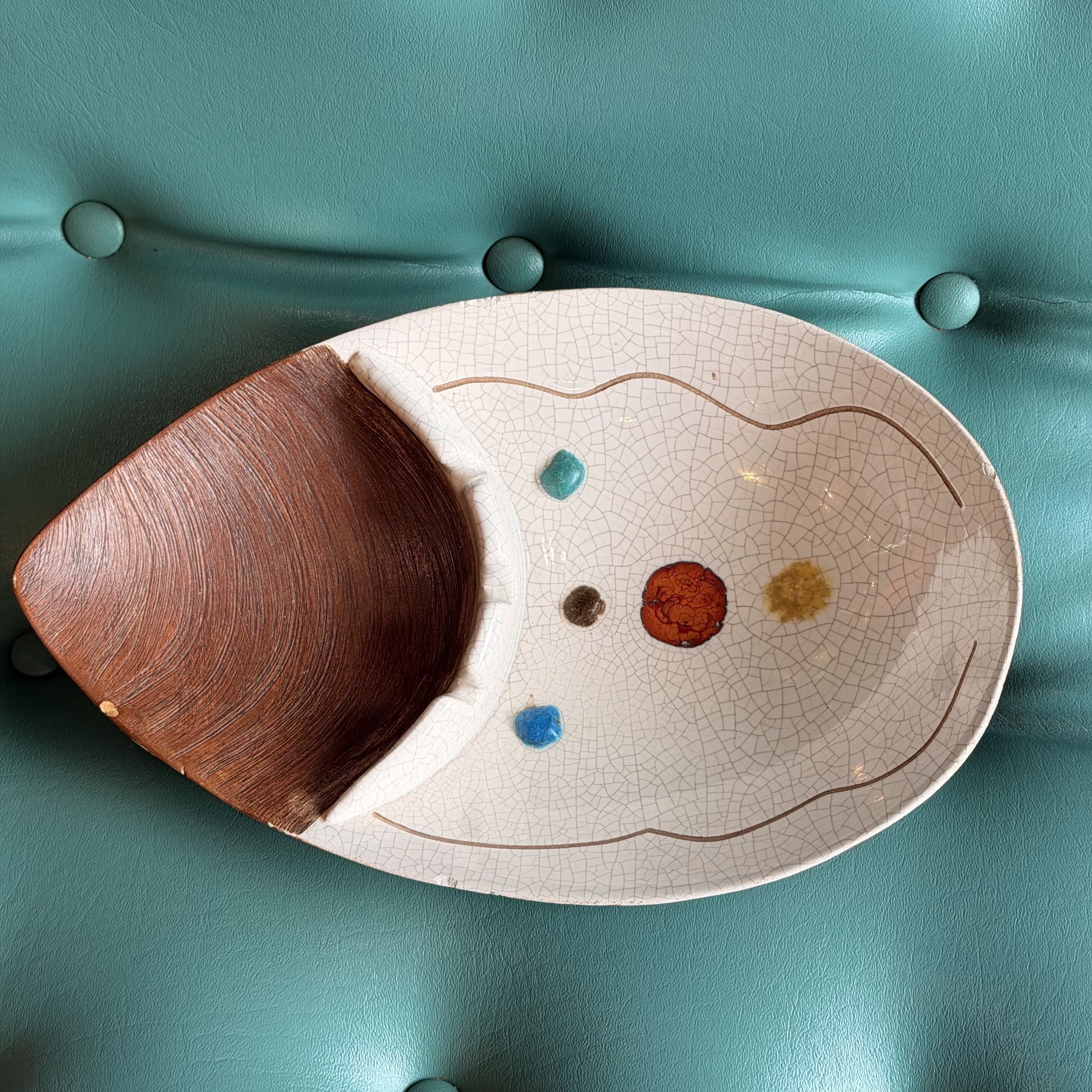 An oval ceramic serving platter with a crackled white glaze and a large brown wooden insert on one side, photographed from above on a teal tufted surface.