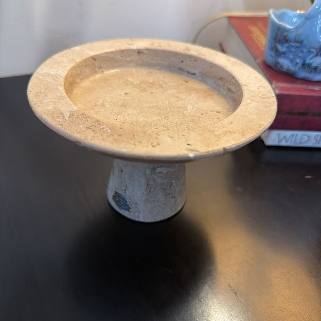 A rustic stone pedestal dish with a wide, shallow round tray on a short cylindrical base, shown on a dark tabletop with a blurred indoor background.