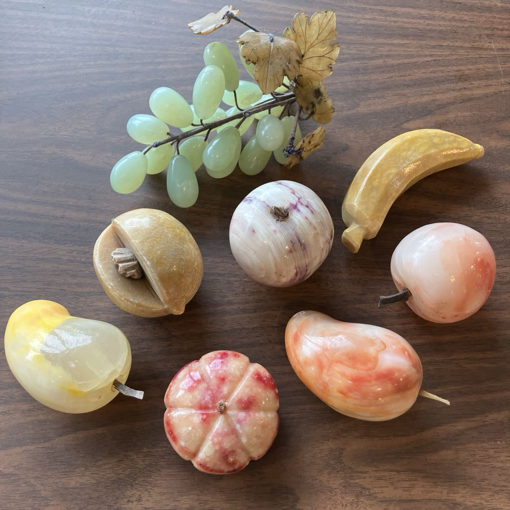 A set of carved stone fruit figurines and a green grape bunch decoration arranged on a dark wood surface.
