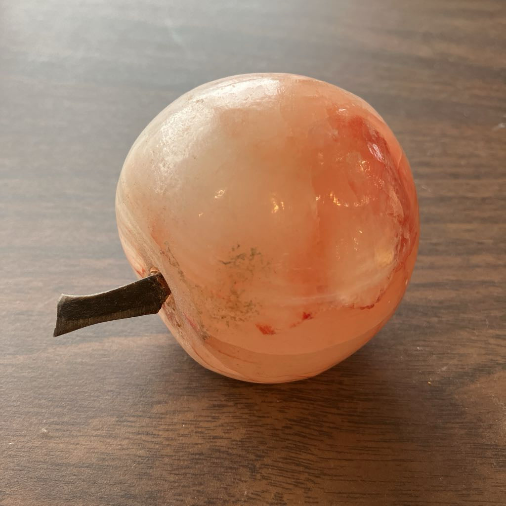 A carved stone fruit sculpture resembling an apple, made from peachy-orange and cream-colored stone with a dark stem, resting on a dark wood surface.