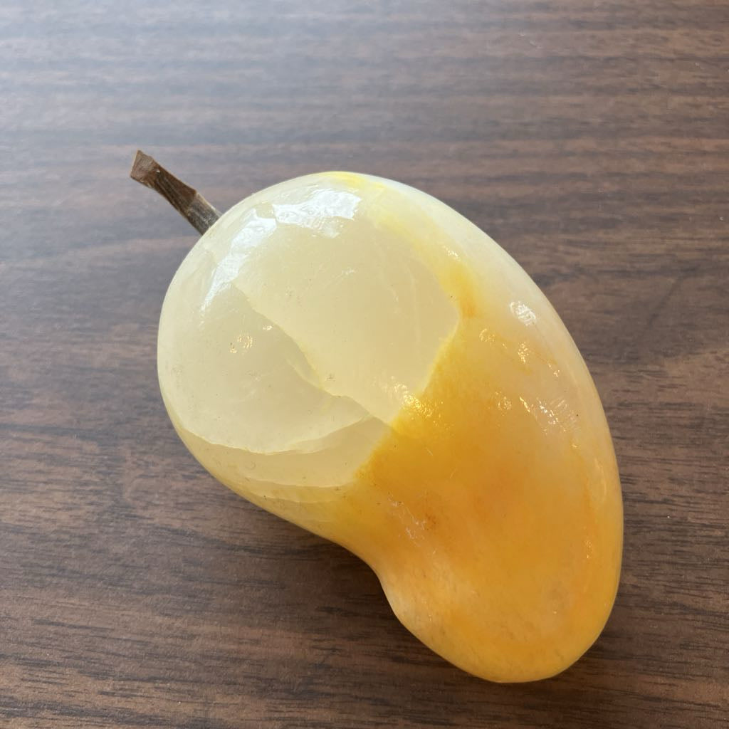 A polished yellow stone sculpture shaped like a mango, featuring a smooth glossy finish and a small brown stem, resting on a dark wood surface.