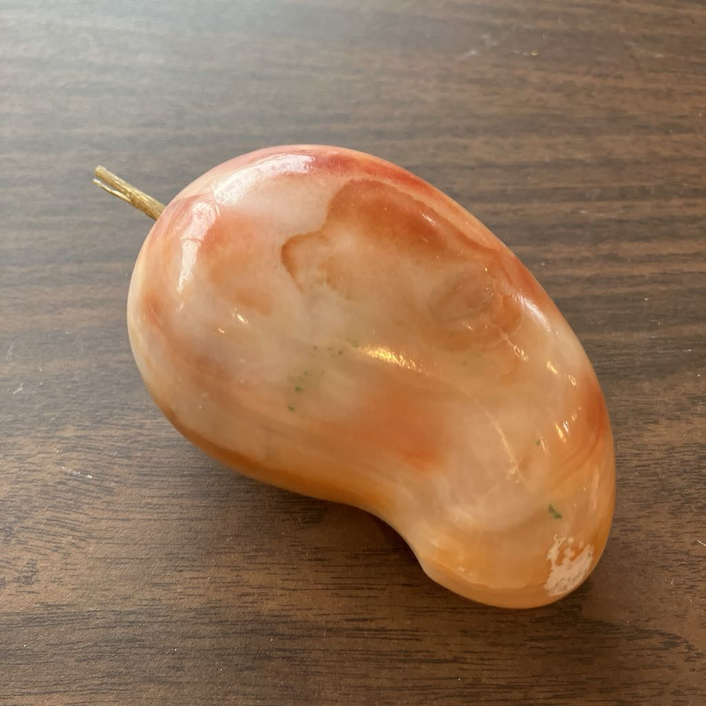 A polished peach and orange stone figurine shaped like a mango with a small gold-toned stem, resting on a dark wood surface.