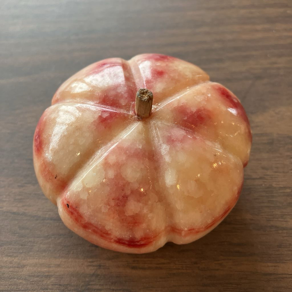 A small decorative pumpkin-shaped object with a glossy finish, featuring a pale peach and cream base with red mottled accents and a short brown stem, placed on a dark wood surface.