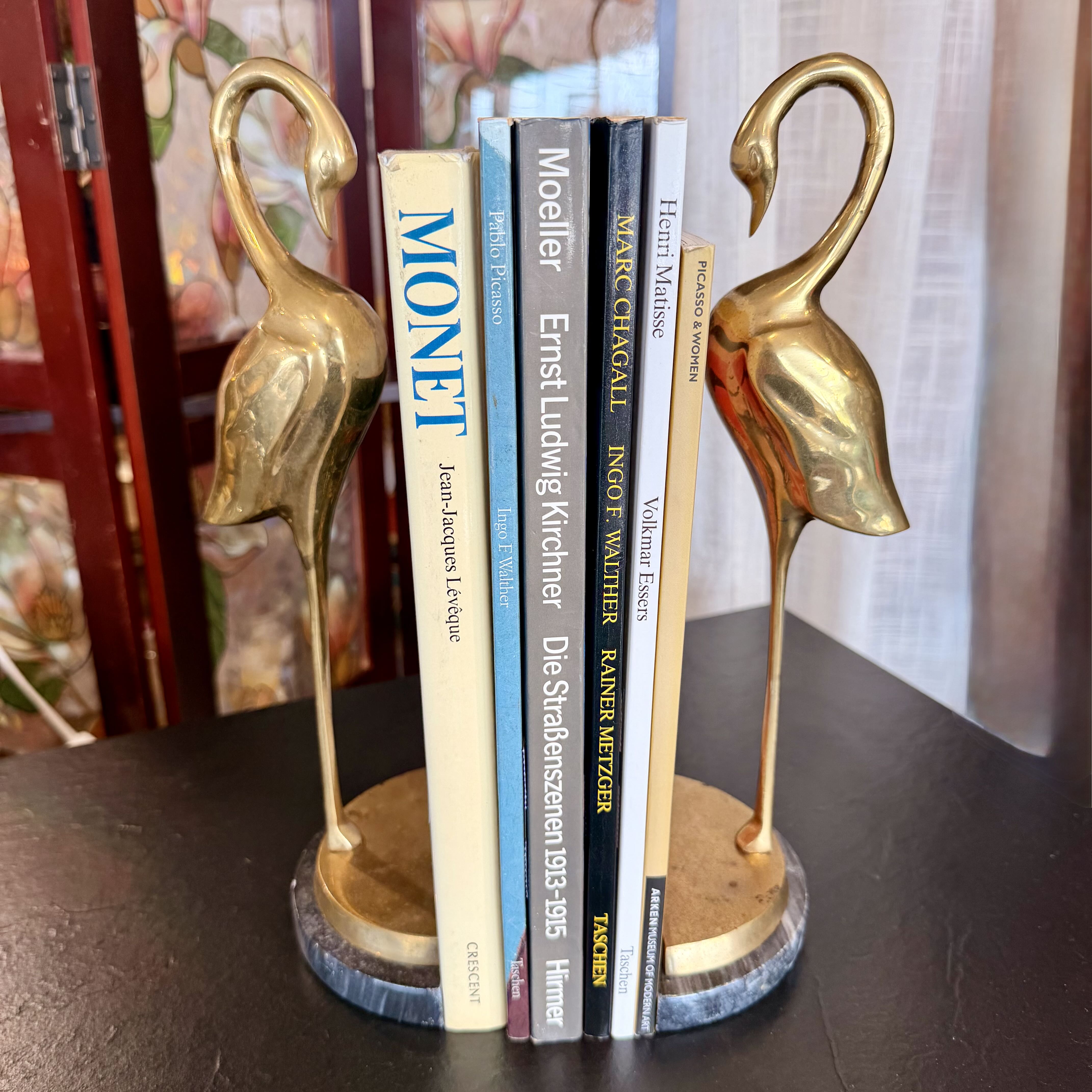 A pair of decorative brass swan bookends holding a row of books on a dark tabletop. The swans have polished gold-toned bodies and stand on round blue-gray marble bases.