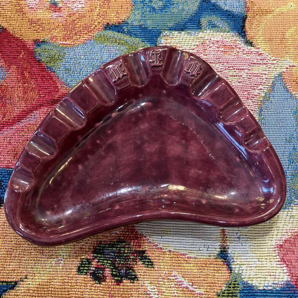 A vintage triangular ceramic dish in a deep burgundy or plum purple color, resting on a colorful floral tapestry.