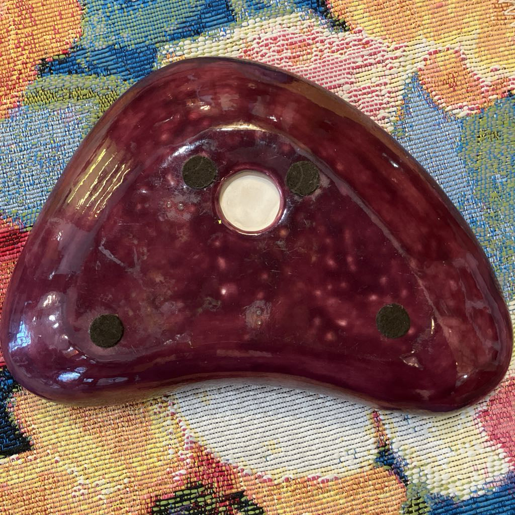 A ceramic dish with a glossy, marbled deep burgundy-red body, featuring a central circular opening and three dark green circular inlays, resting on a colorful patterned textile background.