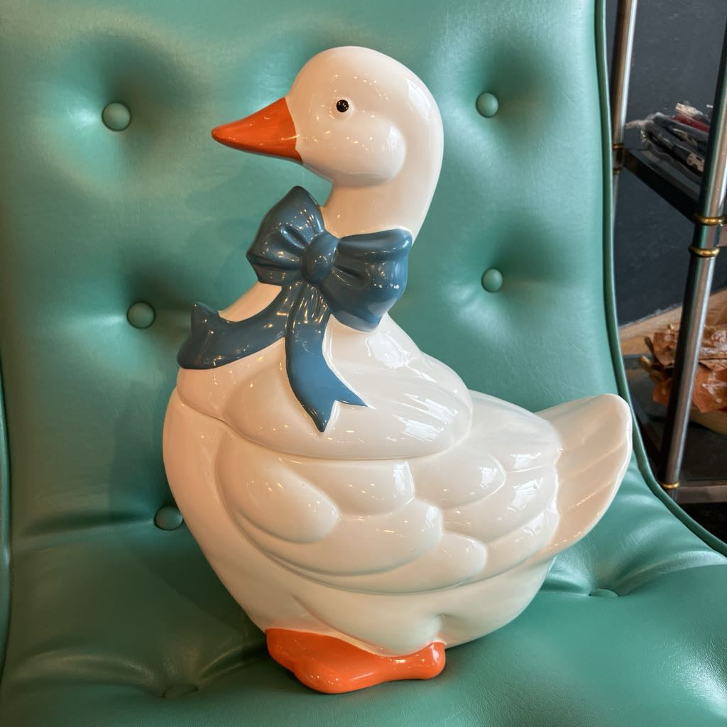 A glossy white ceramic duck figurine with an orange beak and feet, and a dark blue bow around its neck, displayed on a teal tufted chair.