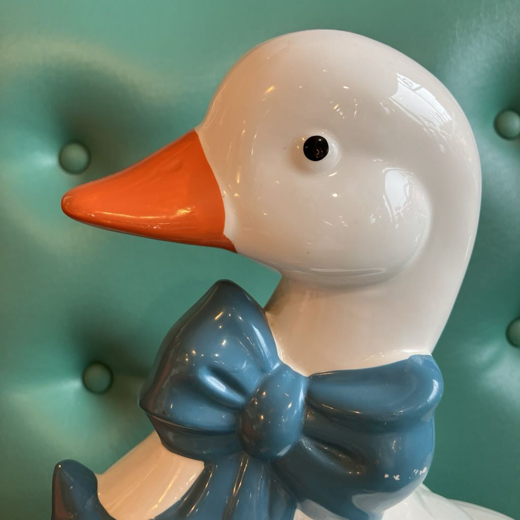 A glossy ceramic duck figurine with a white body, orange beak, and blue bow tie, shown against a teal tufted background.