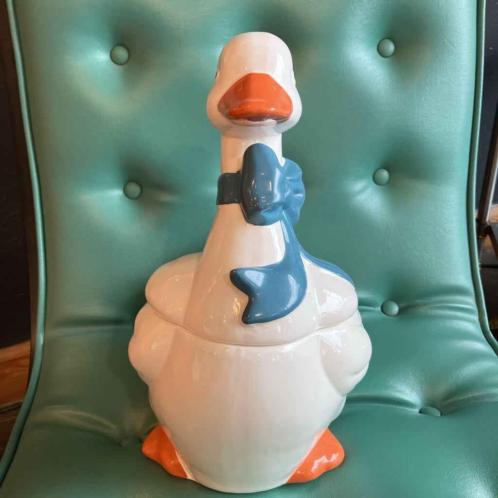 A ceramic duck-shaped container or figurine with a glossy white body, orange beak and feet, and a blue neck bow, displayed on a teal tufted chair.