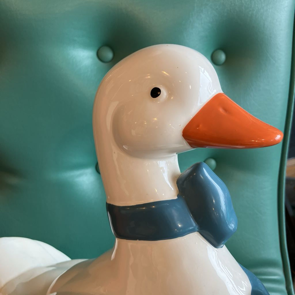 A glossy ceramic duck figurine with a white body, orange beak, and blue bow tie, shown against a teal tufted chair background.
