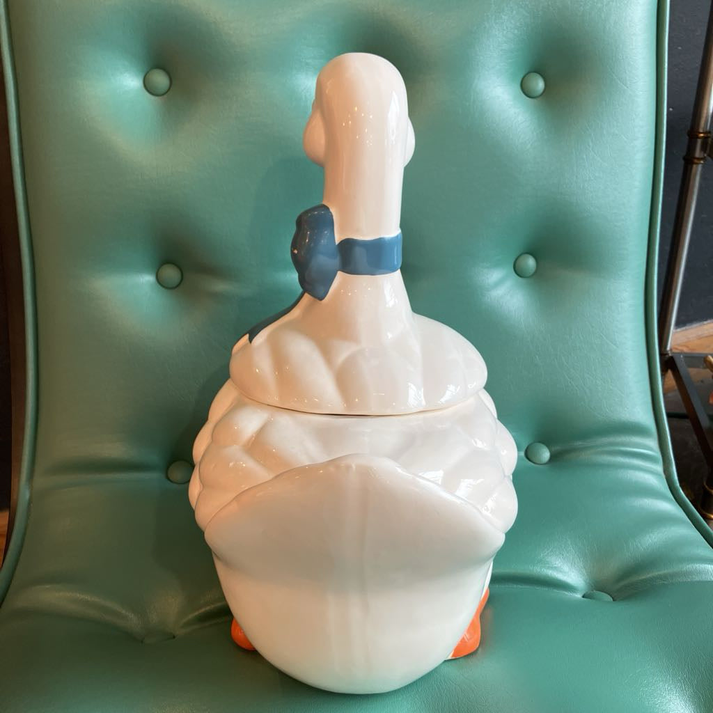 A white ceramic duck-shaped container or tumbler with a blue band around the neck and small orange feet, placed on a teal tufted chair.