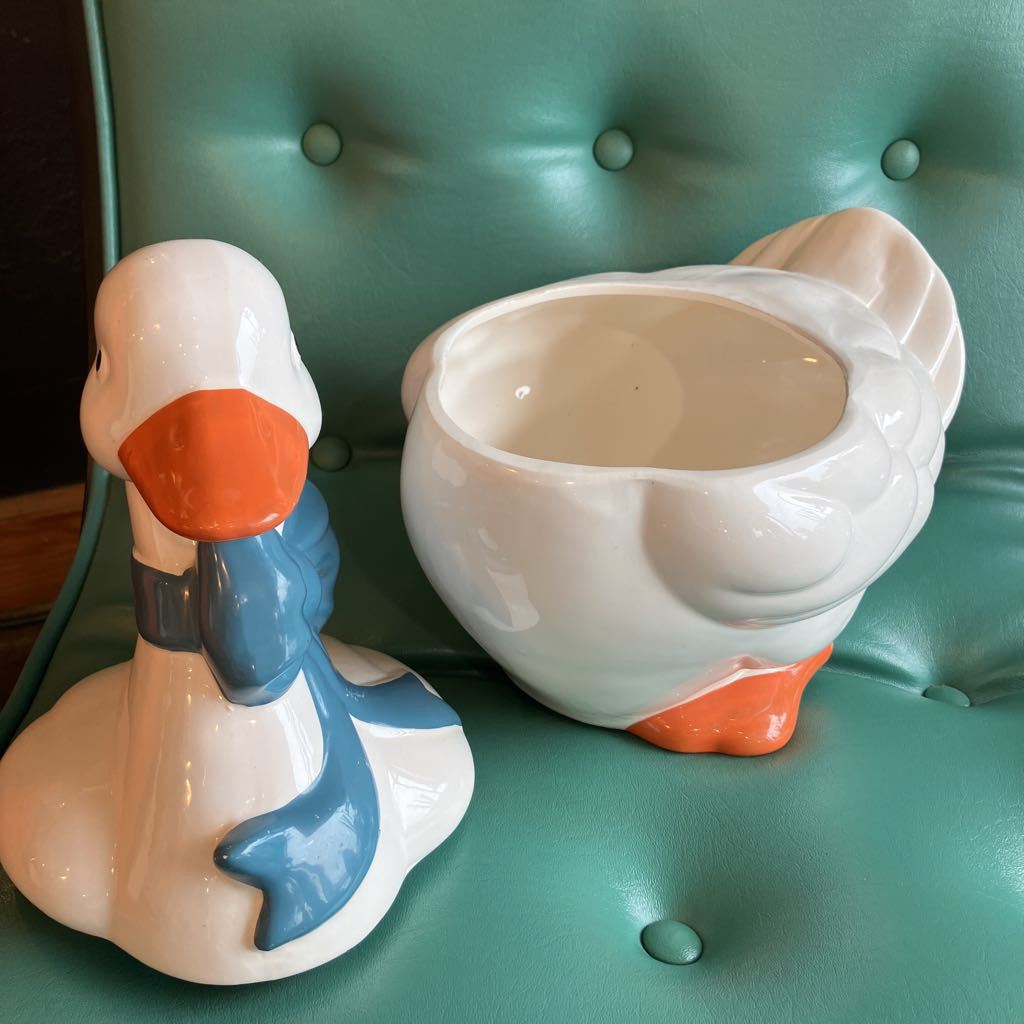 A pair of ceramic duck-shaped decorative pieces, one facing left with an orange beak and blue scarf, and the other turned to show its back with an orange webbed foot, both placed on a teal tufted leather surface.