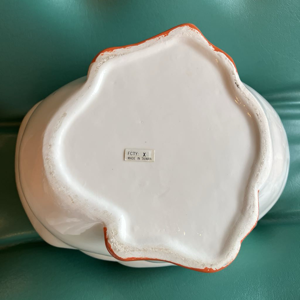 The underside of a white ceramic bowl with a scalloped rim and a thin orange-red edge glaze, resting on a teal-green surface. A small maker's label is visible in the center.