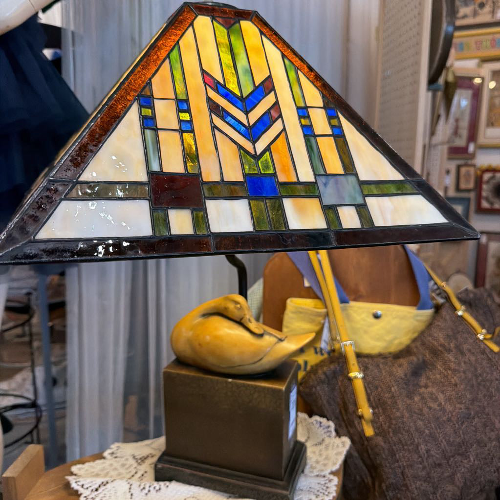A stained glass table lamp with a triangular shade featuring geometric patterns in yellow, green, blue, and white, set on a dark metal base with a sculpted duck figure.