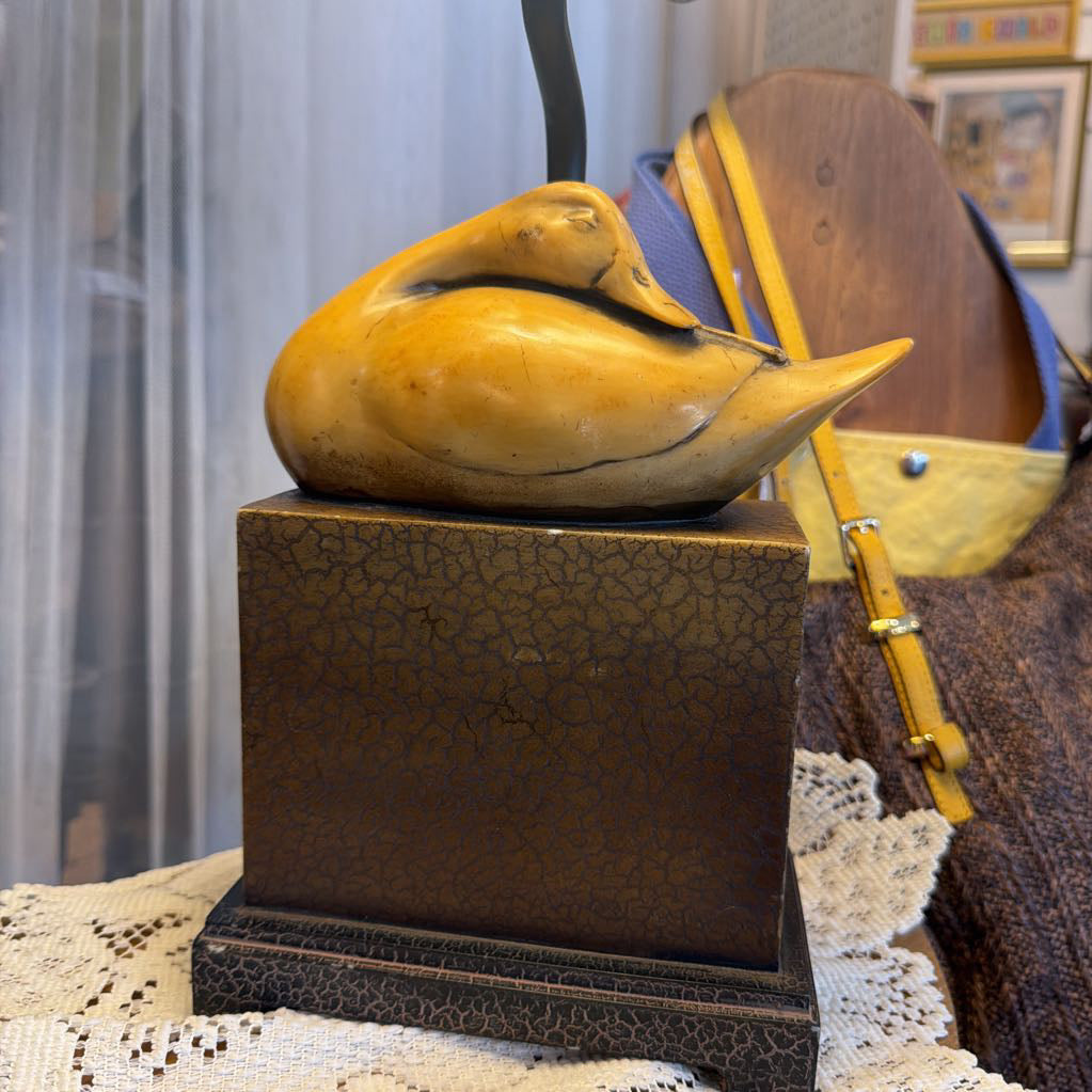 A carved wooden duck sculpture in a natural golden-brown finish, displayed on a dark brown textured square pedestal. The background features a cozy indoor setting with a lace tablecloth, a brown leather bag with yellow straps, and soft curtains.