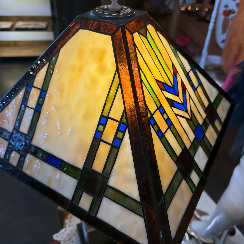 A close-up of a stained glass lampshade with a geometric pattern. The shade features warm yellow and cream glass panels framed by dark brown and black lead lines, with accents of blue, green, and orange.