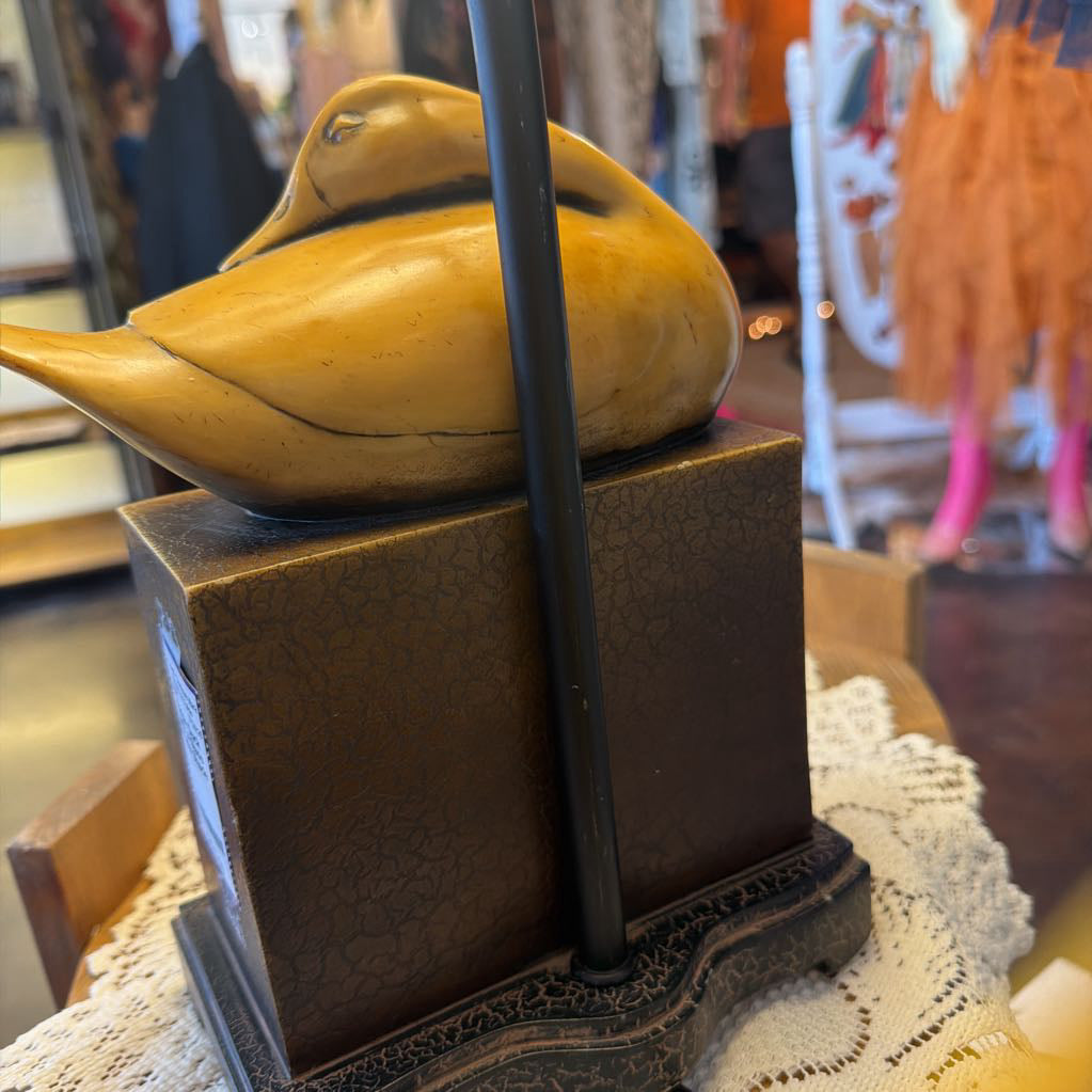 A close-up of a decorative wooden duck sculpture in a warm yellow-brown finish, mounted on a dark textured pedestal with a black vertical pole in front, set in a softly blurred indoor shop environment.