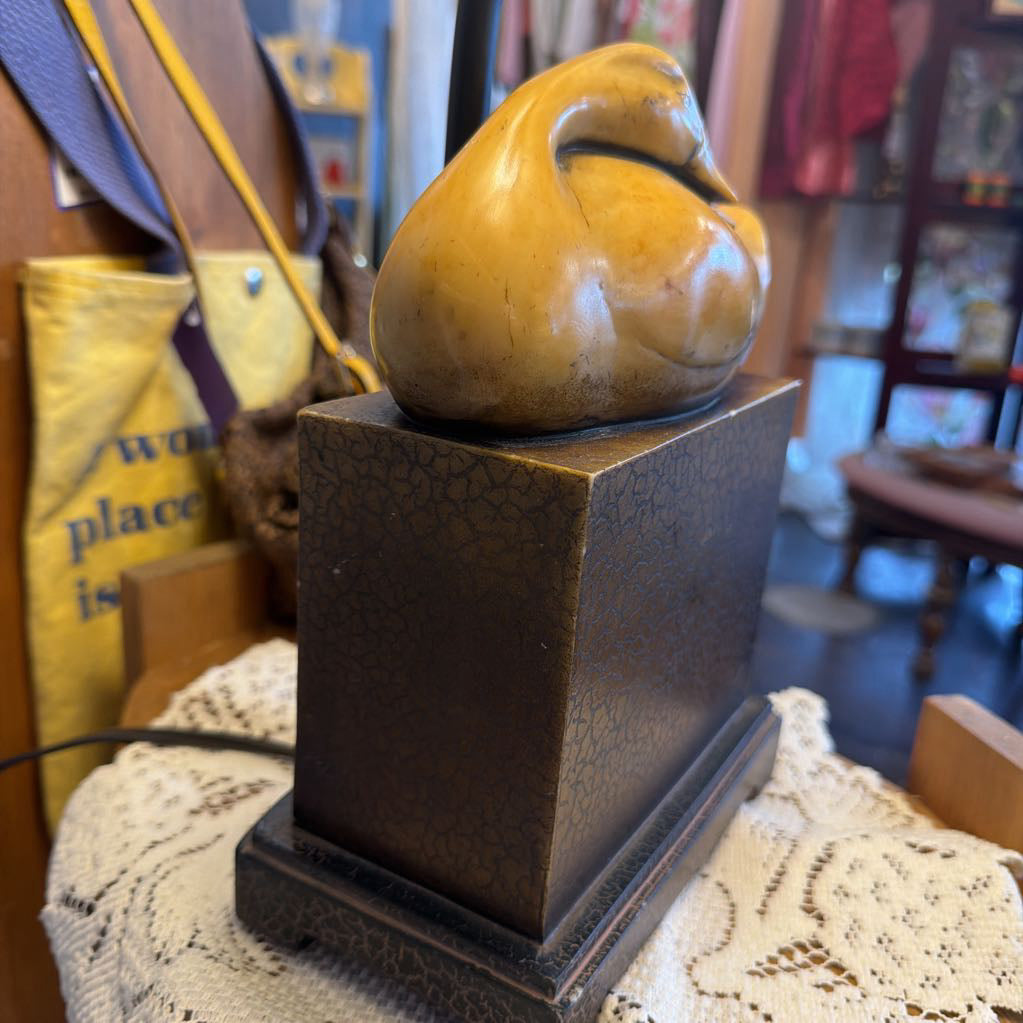 A small sculptural duck figurine in a glossy mustard-yellow finish, mounted on a dark brown square base with a textured finish, set on a white lace doily in a shop.