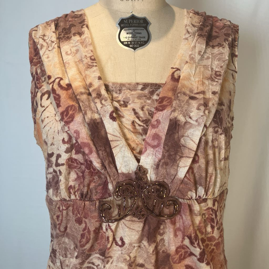 A sleeveless vintage-style dress or tunic with a brown and beige floral pattern, displayed on a cream-colored dress form. The garment features a V-neckline with wide draped shoulder panels and a decorative brown filigree waist detail.