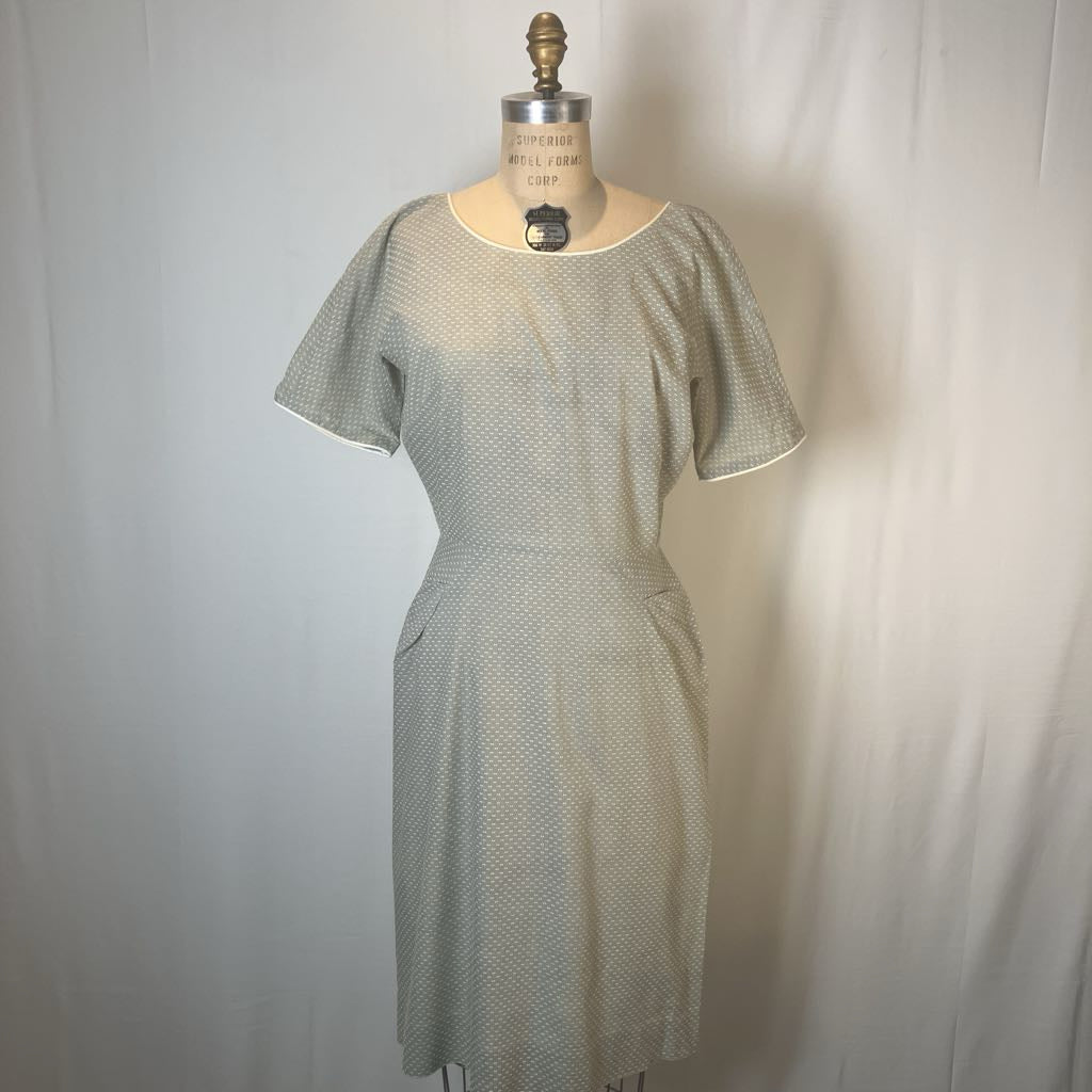 A short-sleeve knee-length dress displayed on a vintage dress form mannequin against a white fabric backdrop. The dress features a fine micro-pattern of small green and white dots, with solid white piped edges on the short sleeves and neckline.