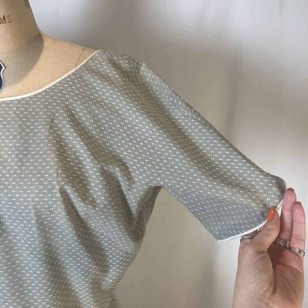 A close-up of a short-sleeve top on a dress form, featuring a small white geometric pattern on a gray base. A hand is holding the sleeve to show its length and cuff detail.