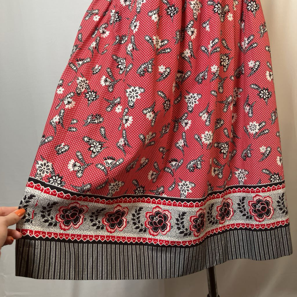 A red skirt with a white and black floral pattern and a decorative border, held up by a hand against a plain light background.
