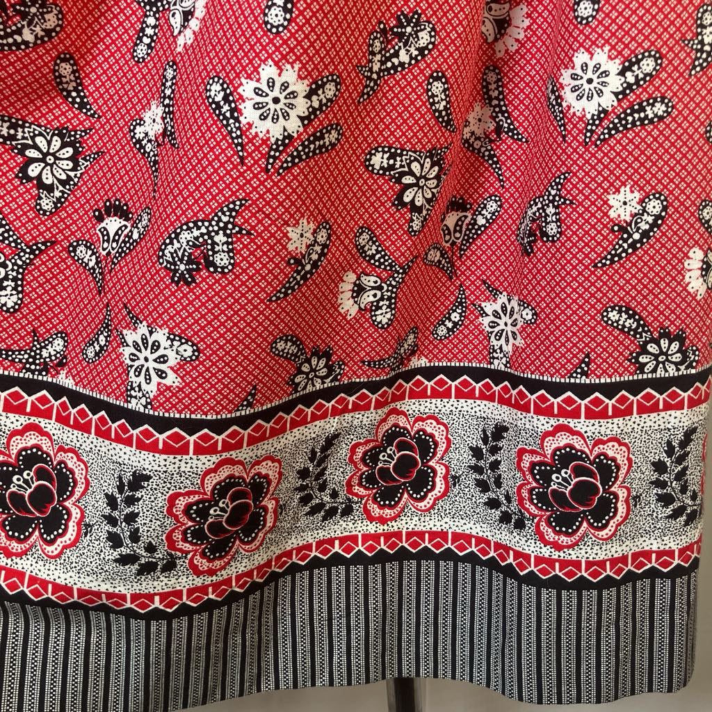 A close-up of a red patterned fabric with black and white floral motifs, a decorative border band, and a striped hem.