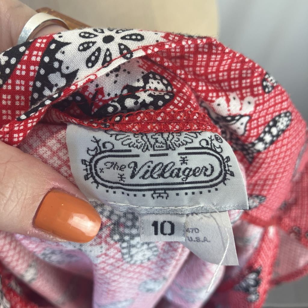 A close-up of a patterned fabric garment held by a hand with orange nail polish, showing a sewn-in label that reads 'The Villagers' and size '10'. The fabric has a red, pink, white, and black floral and geometric pattern.