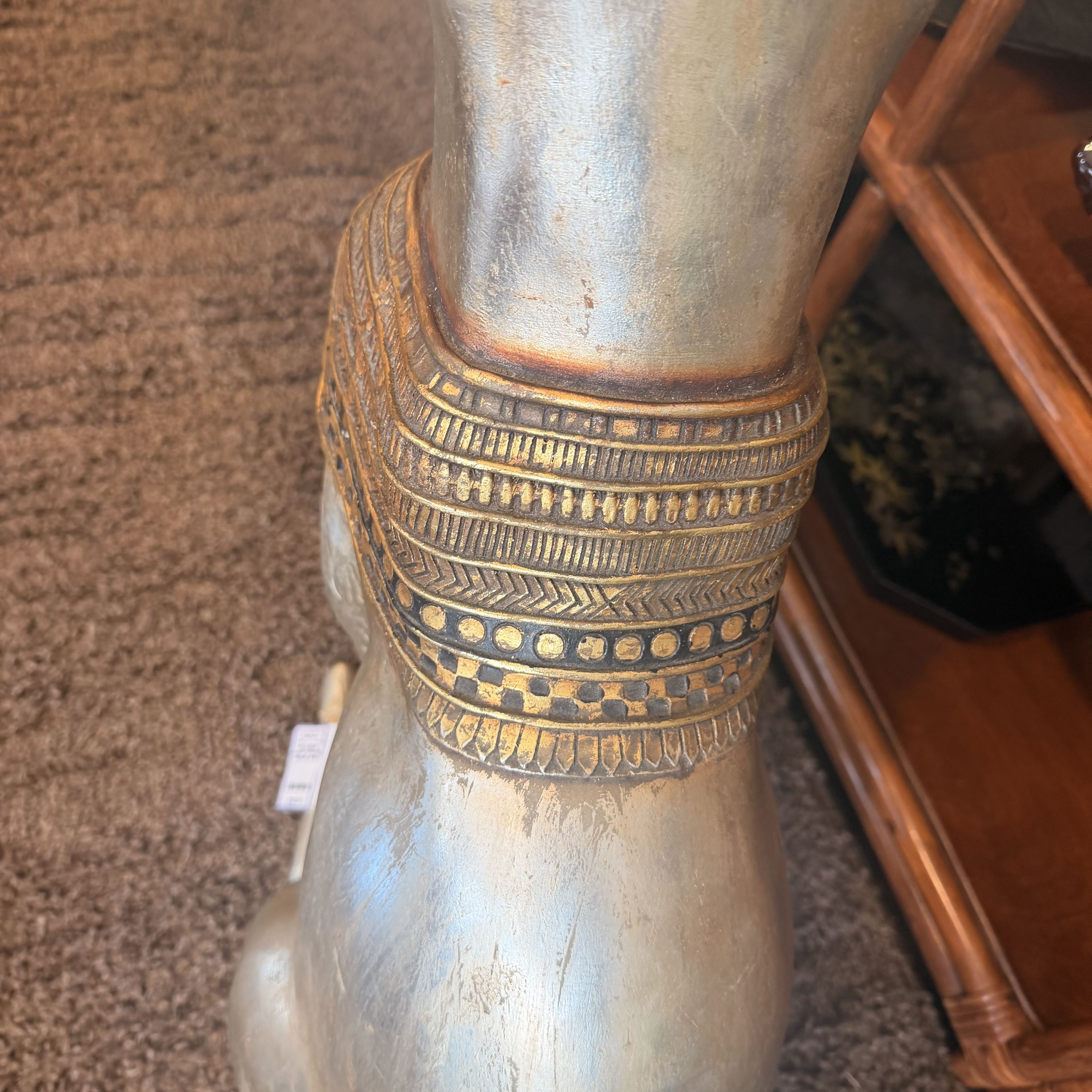 A close-up of a metallic silver decorative stand or pedestal with a wide gold-toned patterned band around the upper section, photographed indoors on a carpeted floor next to a wooden table.