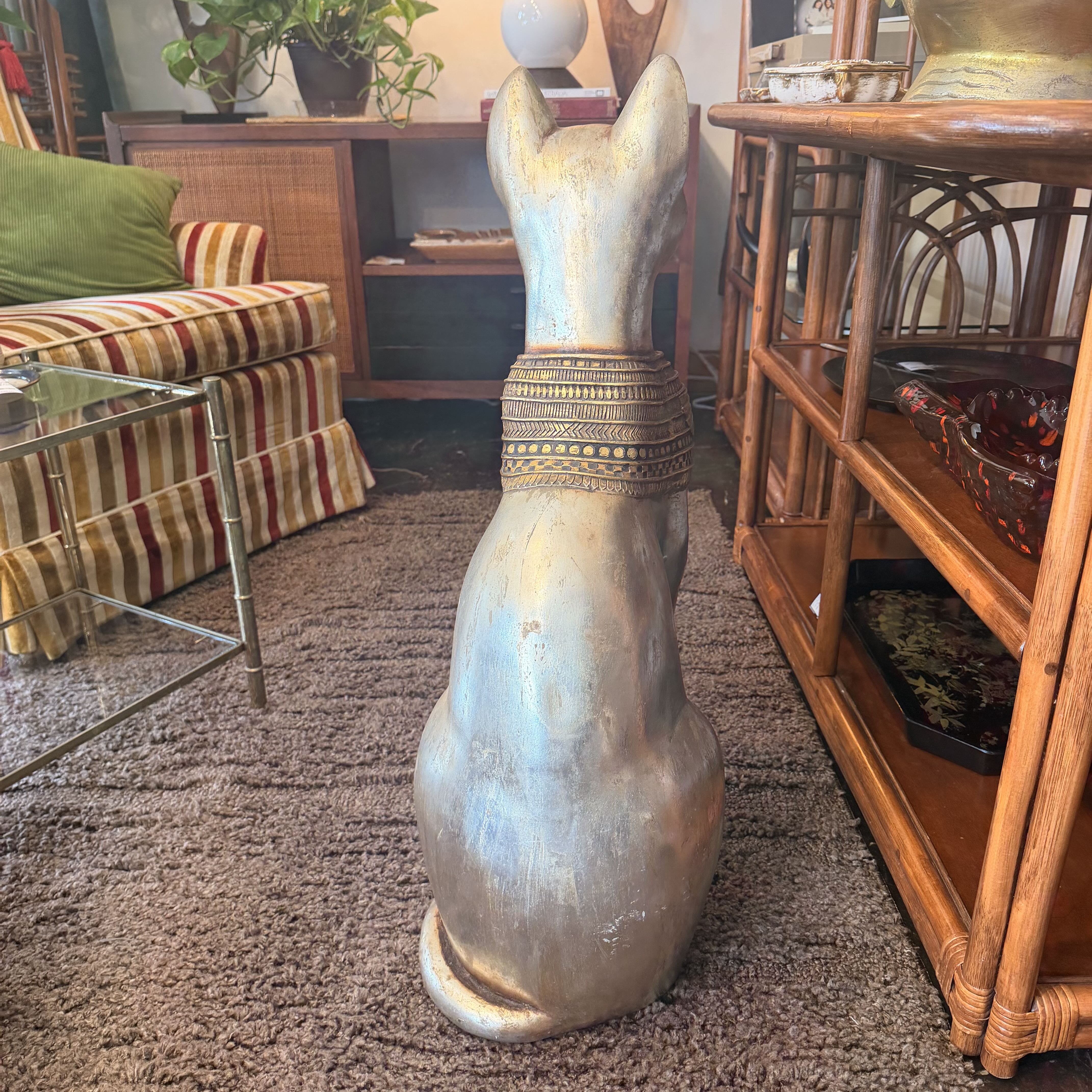 A tall, seated cat figurine or decorative sculpture in a distressed silver finish with a gold patterned collar, photographed indoors on a carpeted floor between furniture pieces.