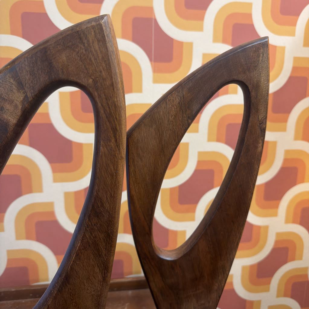 Close-up of two dark brown wooden chair backs with curved, looped cutouts, set against a retro patterned wallpaper background in orange, yellow, and red.