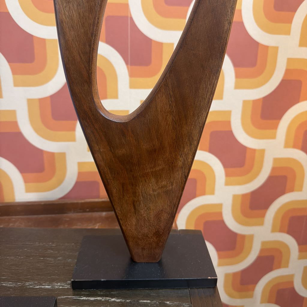 A modern abstract wooden sculpture with a dark brown wood grain finish, mounted on a black rectangular base. The background features a retro patterned wallpaper in warm tones of orange, yellow, and red.