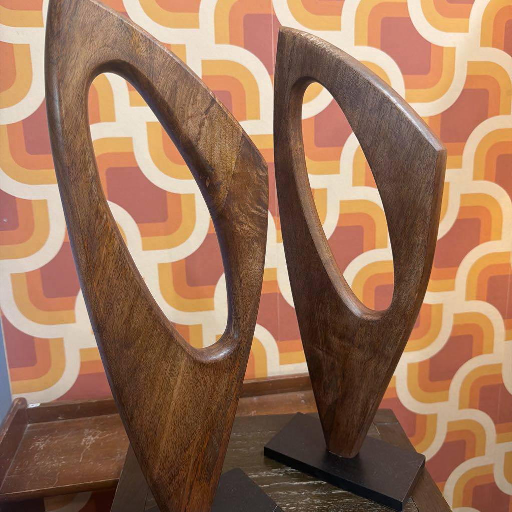 Two abstract wooden wall art sculptures with curved, looped shapes mounted on black square bases, set against a retro patterned wallpaper background.