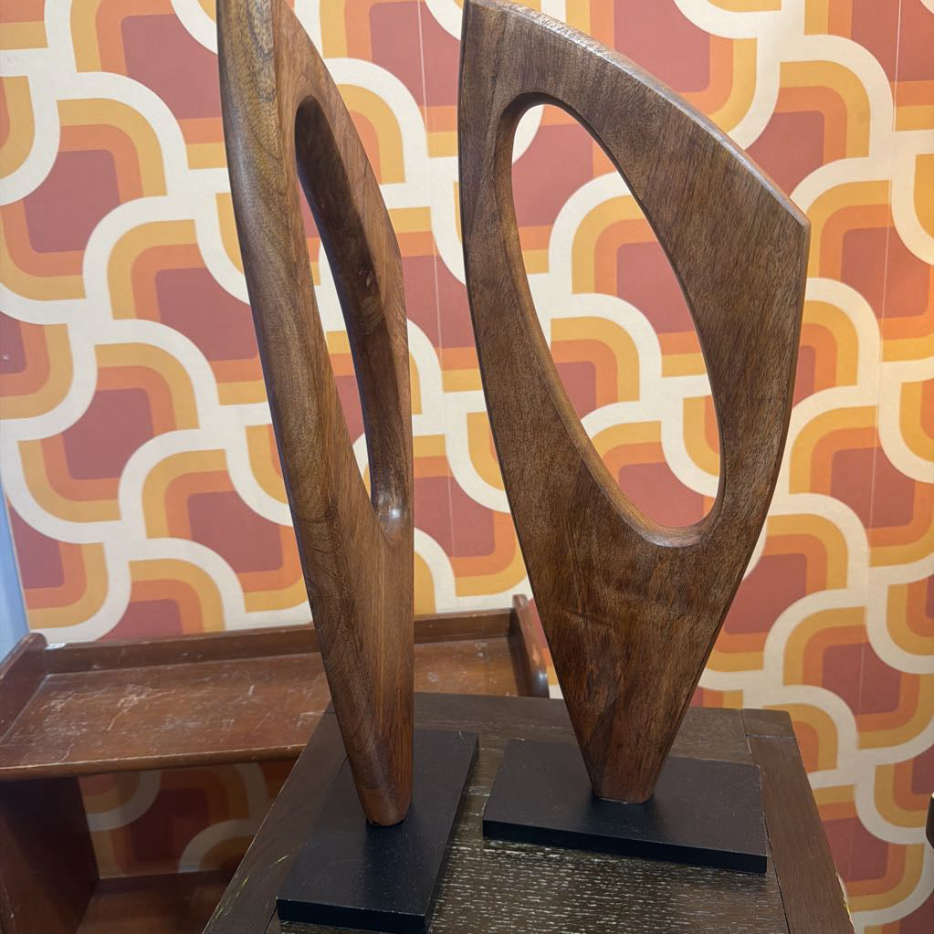 Two mid-century modern wooden abstract sculptures with hollow geometric shapes, displayed on black bases against a retro patterned wallpaper background.
