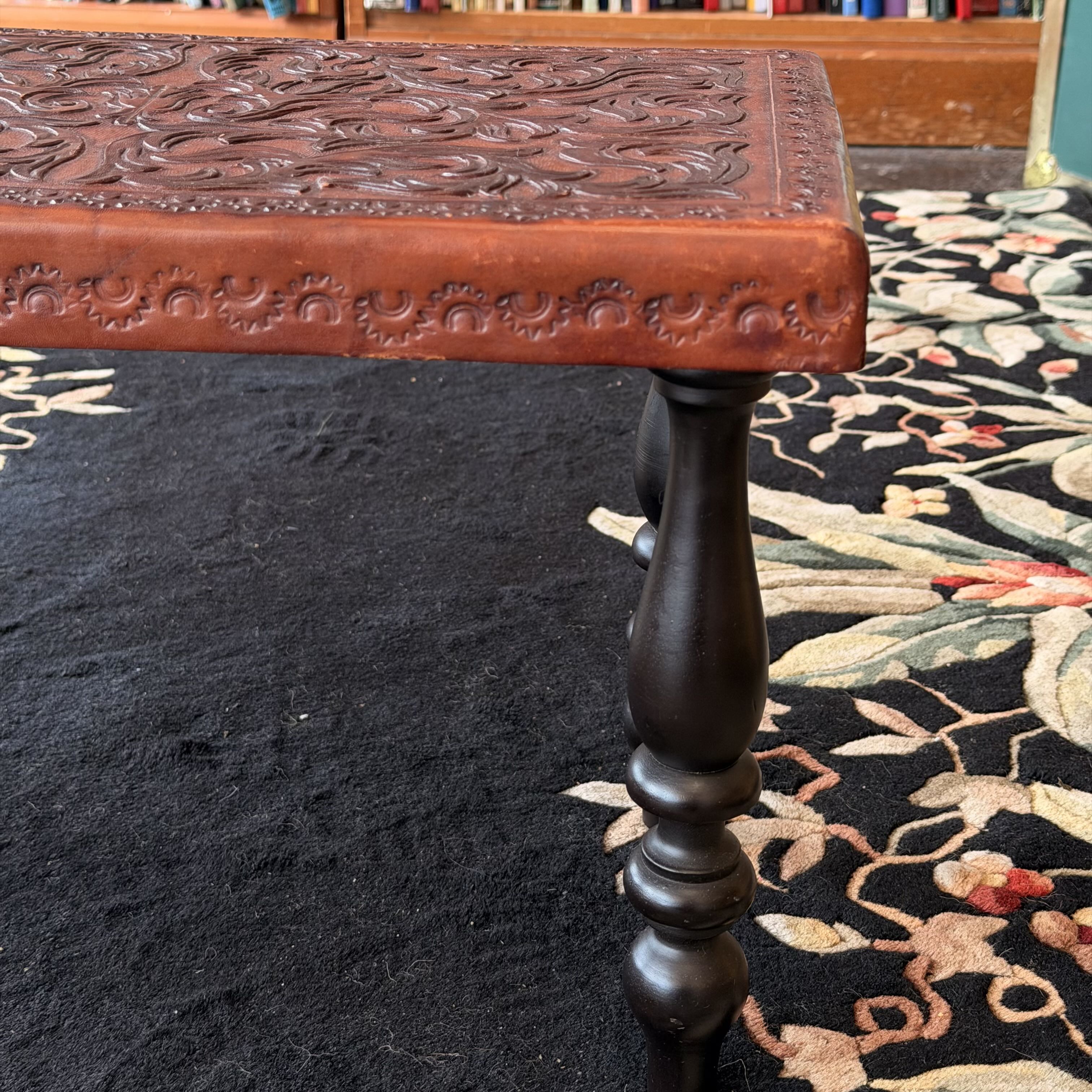 Close-up of a carved wooden table corner with an ornate reddish-brown top and a dark turned leg, placed on a black floral area rug in an indoor room.