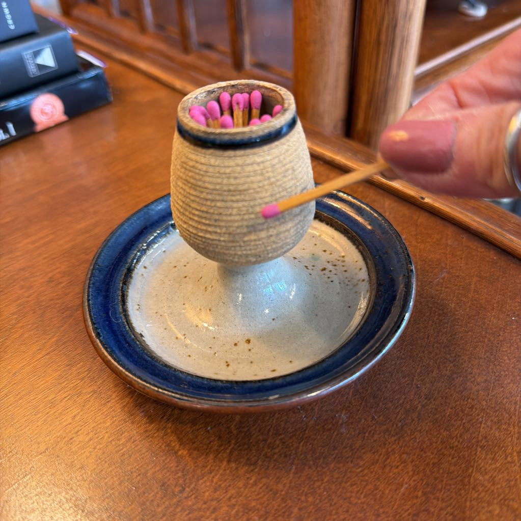 A ceramic match holder shaped like a small vase, filled with pink-tipped matches, sits on a matching round saucer on a wooden table. A hand is holding a match near the top.