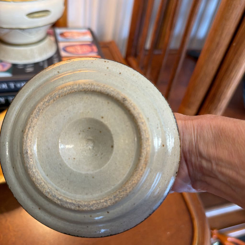 A close-up of a ceramic bowl or cup base held by a hand, showing a speckled off-white glaze and a dark rim, with a wooden chair and books in the background.