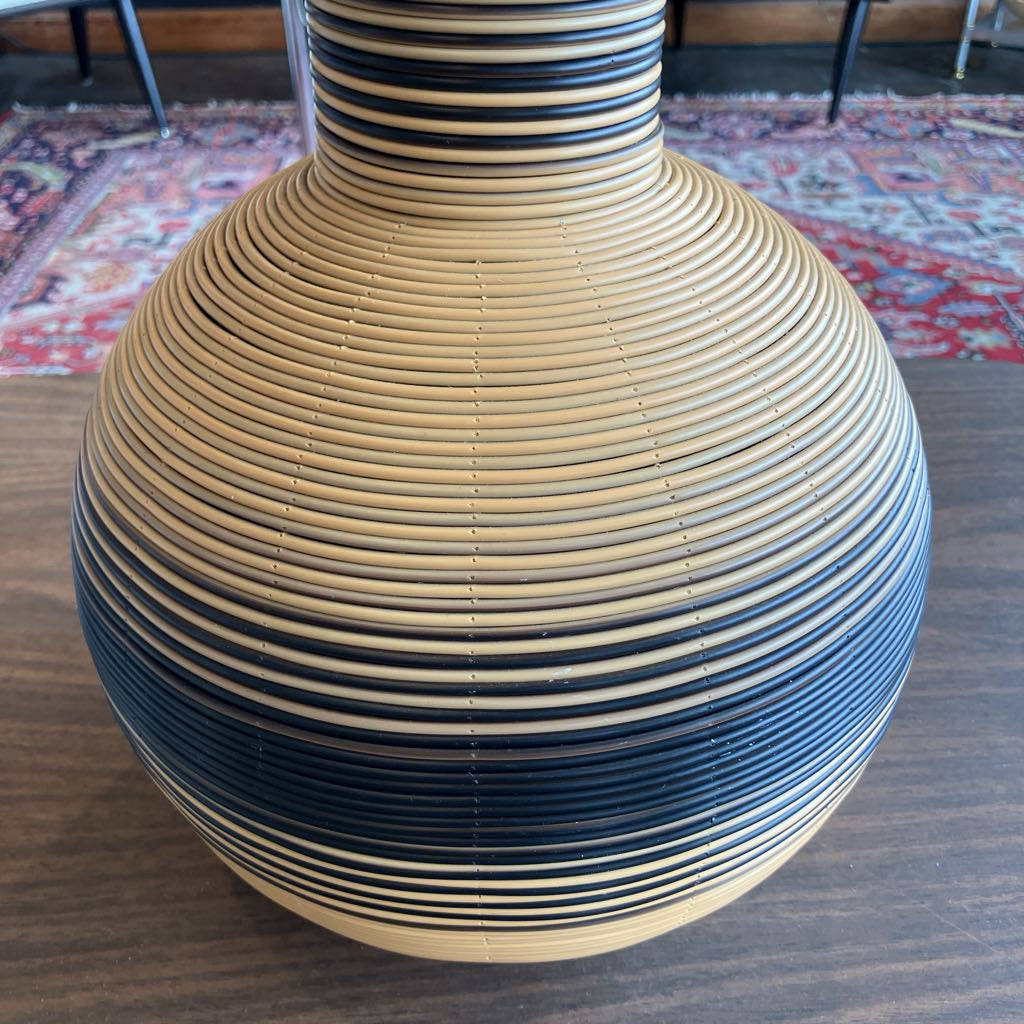 A large round vase with a narrow neck, featuring horizontal woven stripes in natural beige and dark navy blue, sitting on a wooden table with a patterned rug in the background.