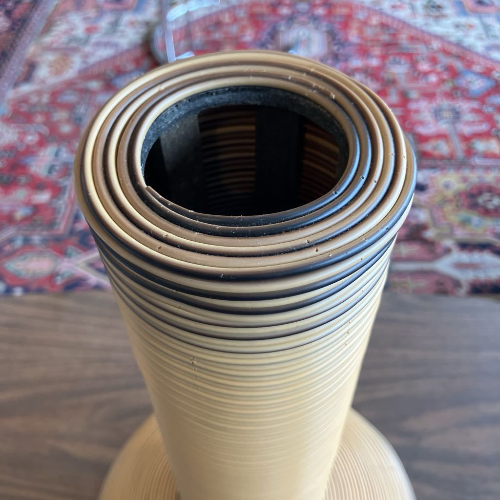 A close-up of a beige and black striped coiled column or decorative object, photographed on a wooden surface with a patterned rug in the background.