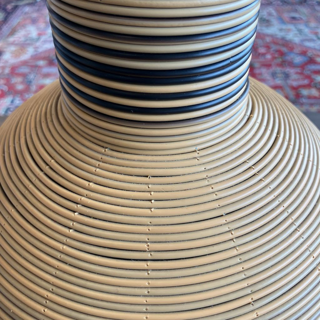 A close-up of a woven rattan or wicker planter with a flared beige body and a narrow neck featuring alternating beige and dark blue horizontal stripes, set against a blurred patterned rug background.